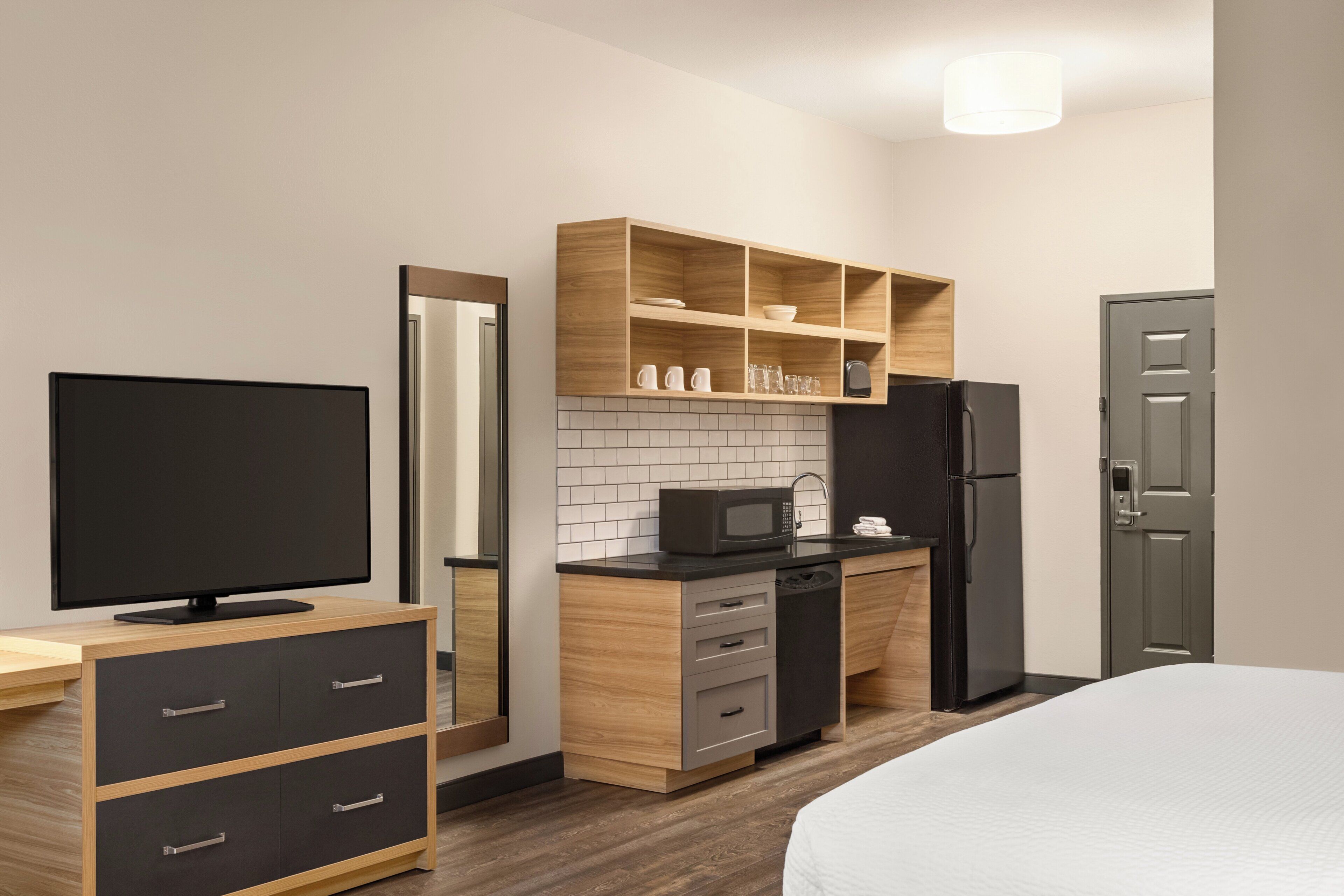 A modern hotel room with a flat-screen TV, a wooden cabinet with drawers, a microwave, a refrigerator, and a bed.