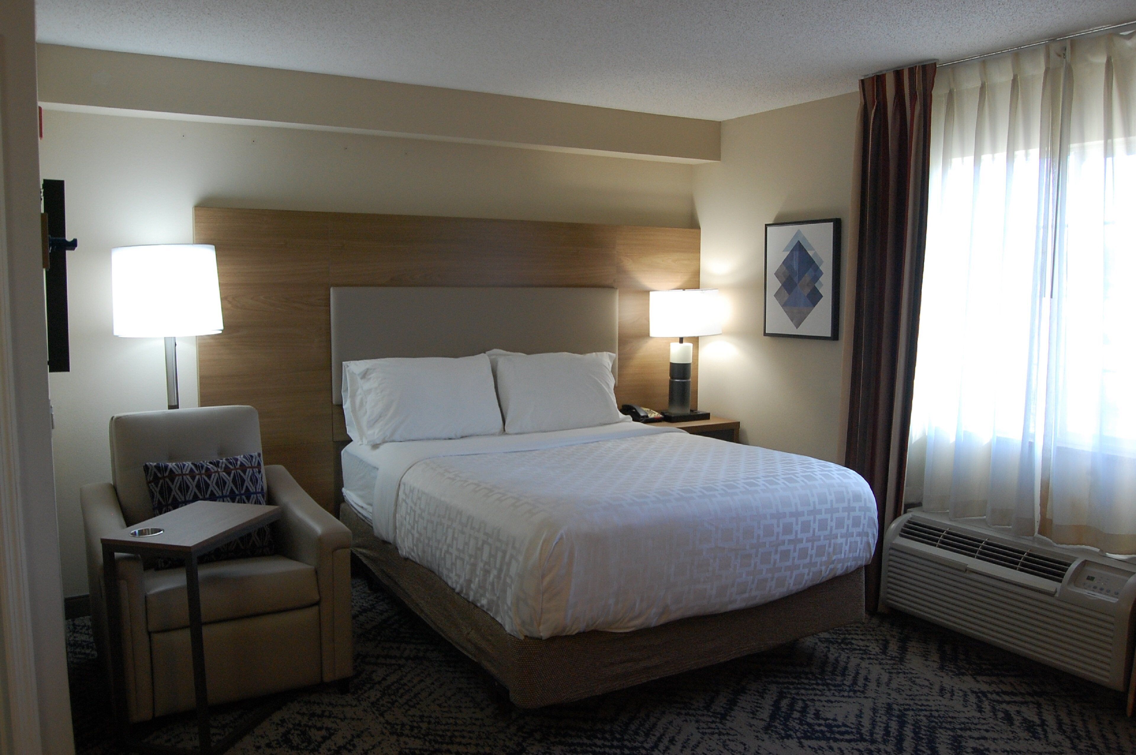 A hotel room with a bed, a chair, a desk, a nightstand, a lamp, a picture on the wall, and an air conditioning unit.