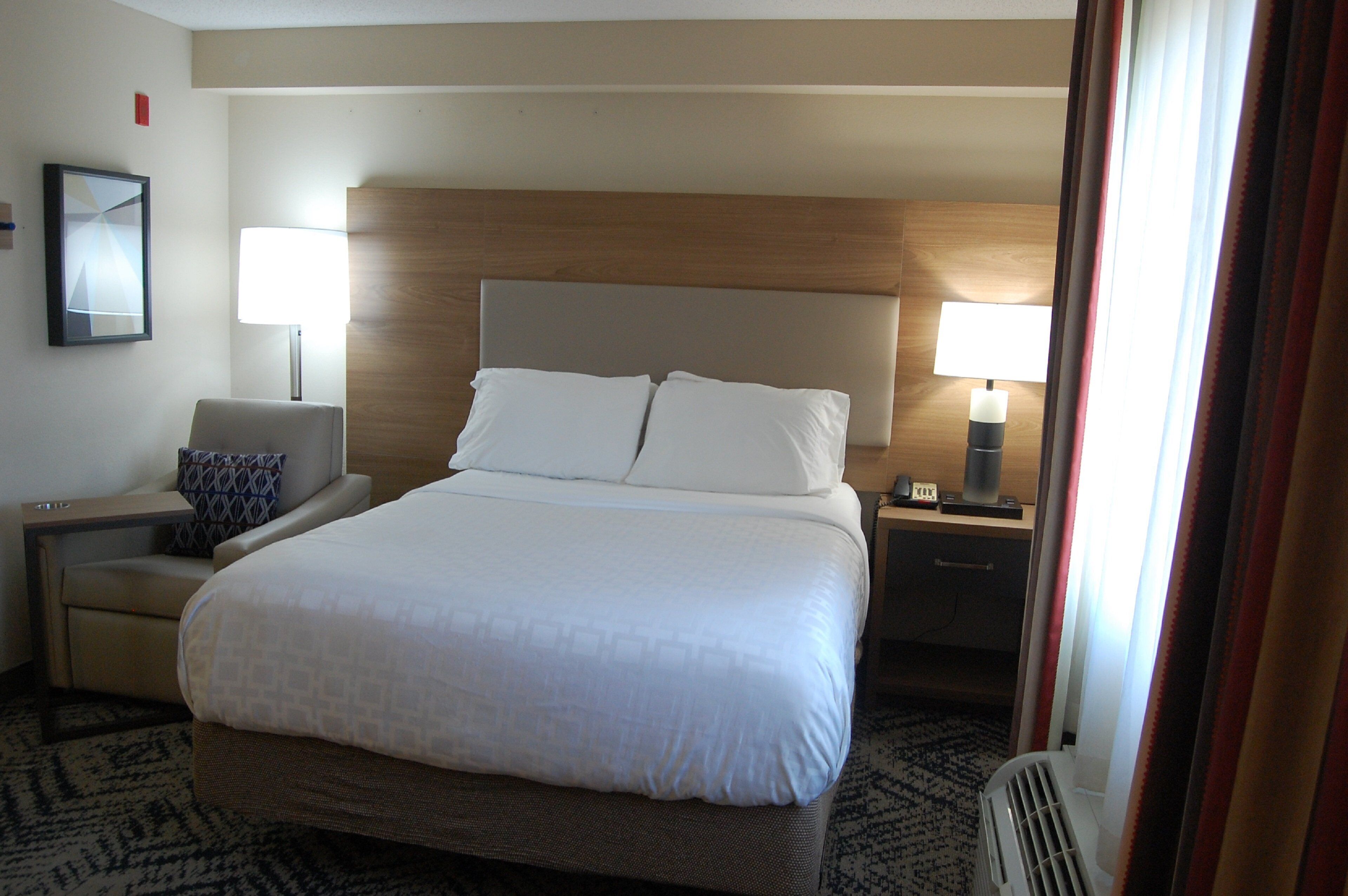A hotel room with a large bed, a bedside table with a lamp, a chair, and a small table with a coffee maker.
