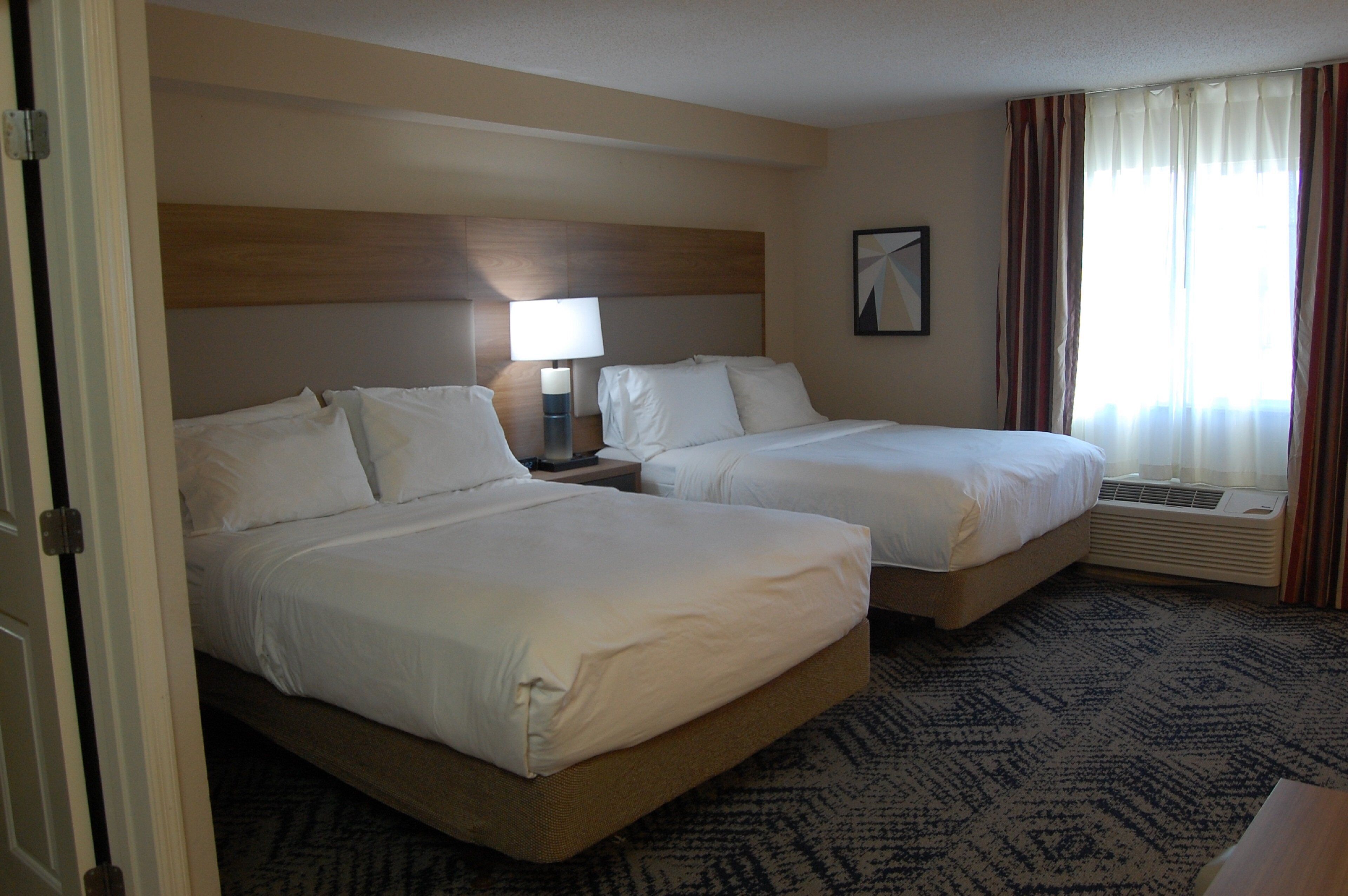 A hotel room with two beds, a nightstand with a lamp, a window with curtains, and a carpeted floor.
