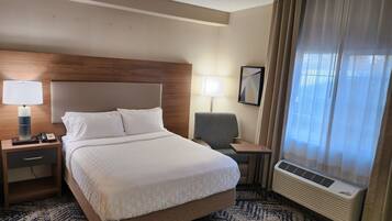 A hotel room with a bed, bedside tables, a lamp, a chair, a mirror, and an air conditioning unit.