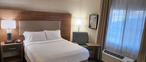 A hotel room with a bed, bedside tables, a lamp, a chair, a mirror, and an air conditioning unit.