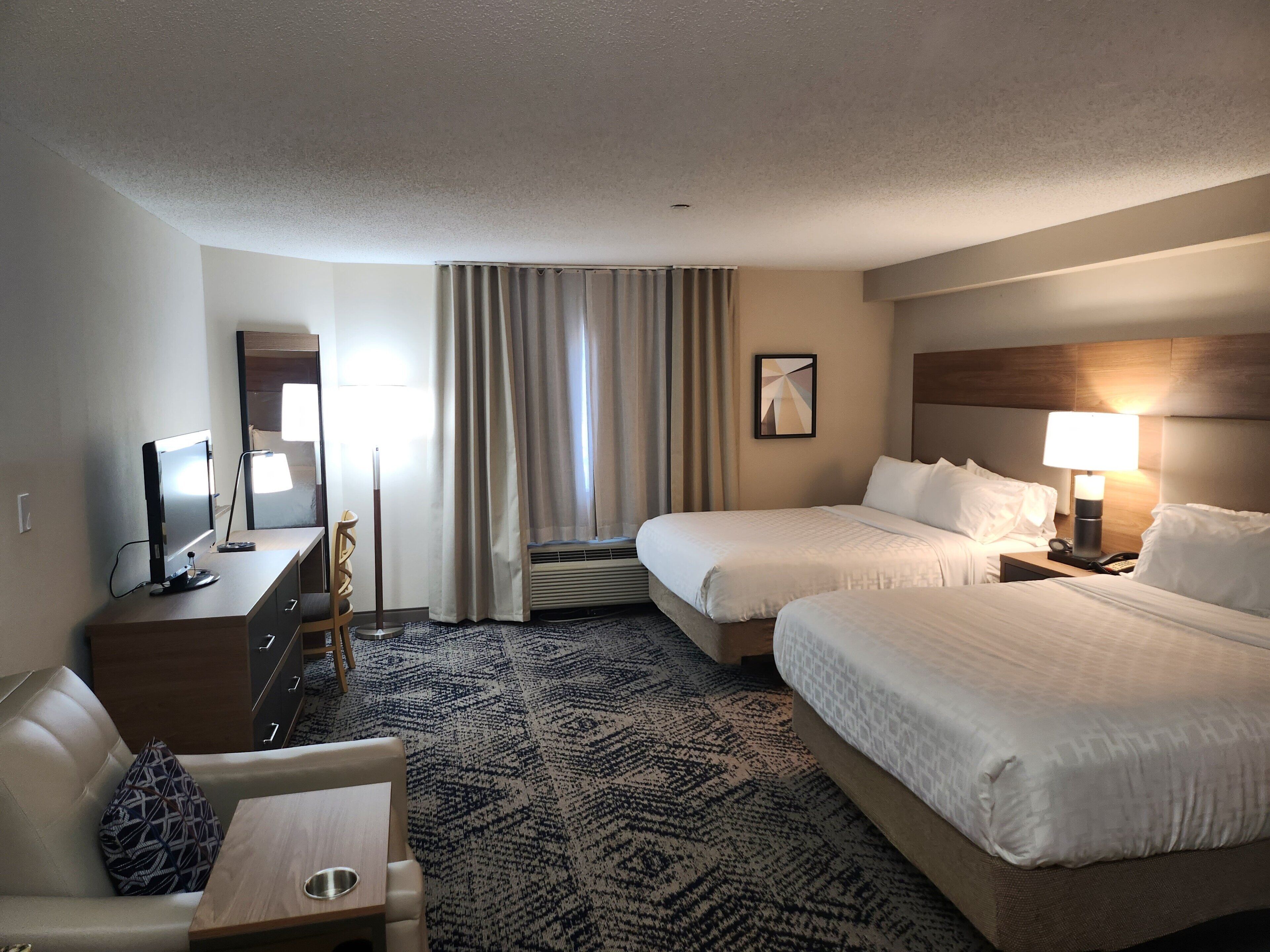 Habitación de hotel con dos camas, un escritorio con computadora, una silla, una lámpara y un ventanal con cortinas.