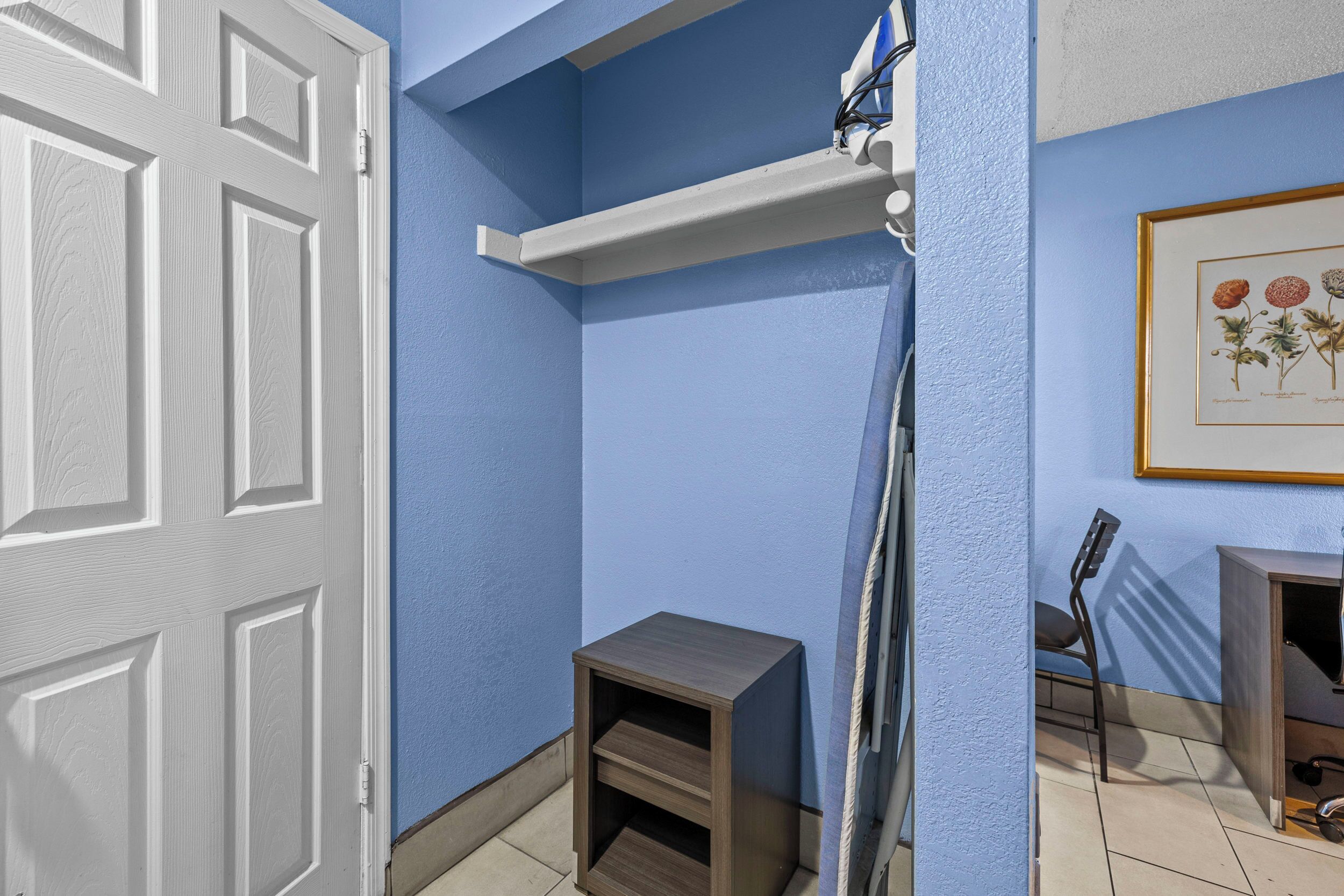 Une pièce avec un mur bleu, une porte blanche, un petit meuble en bois, une étagère blanche, une illustration botanique encadrée et une chaise.