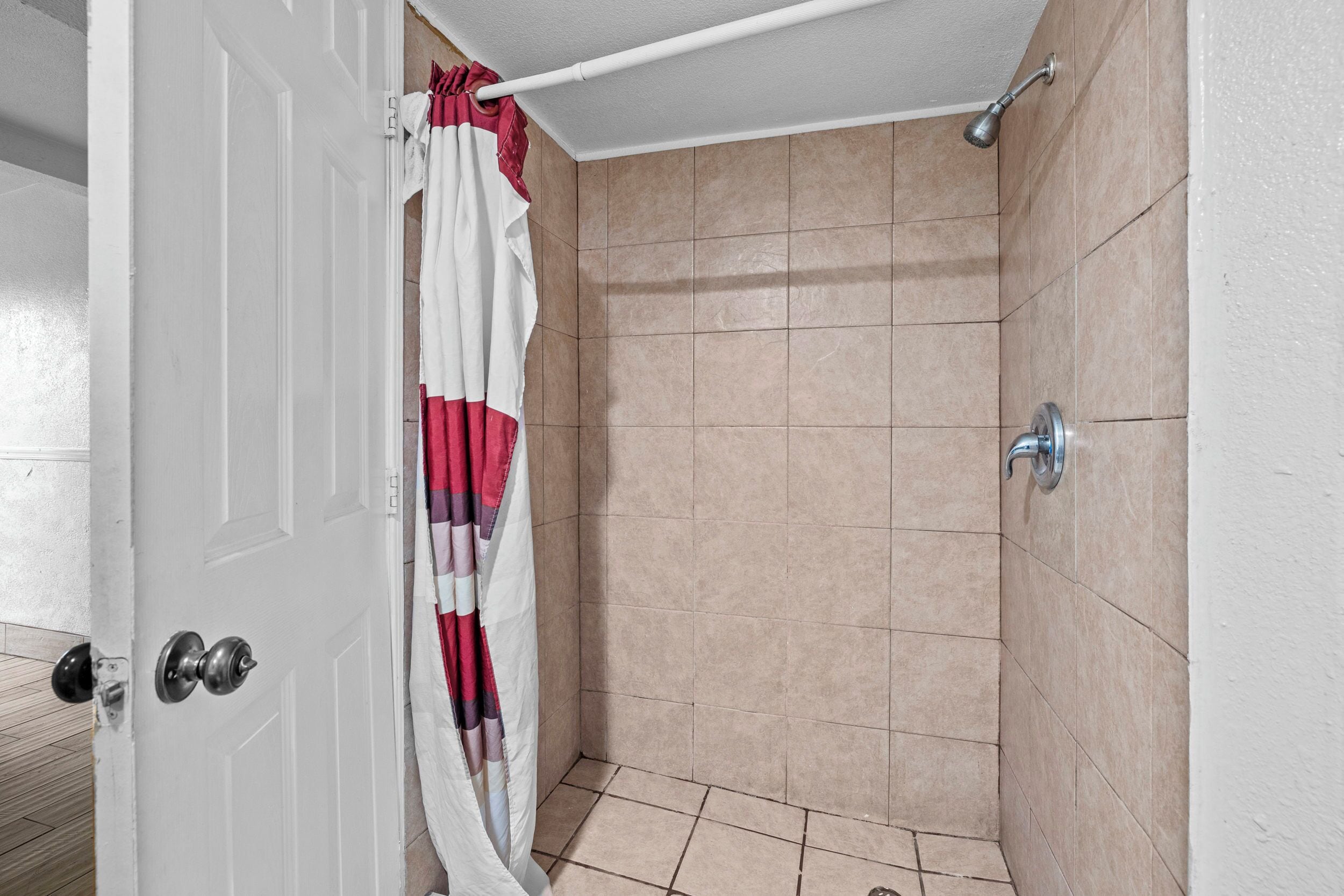 A small, enclosed shower area with beige tiles, a white door, and a shower curtain.
