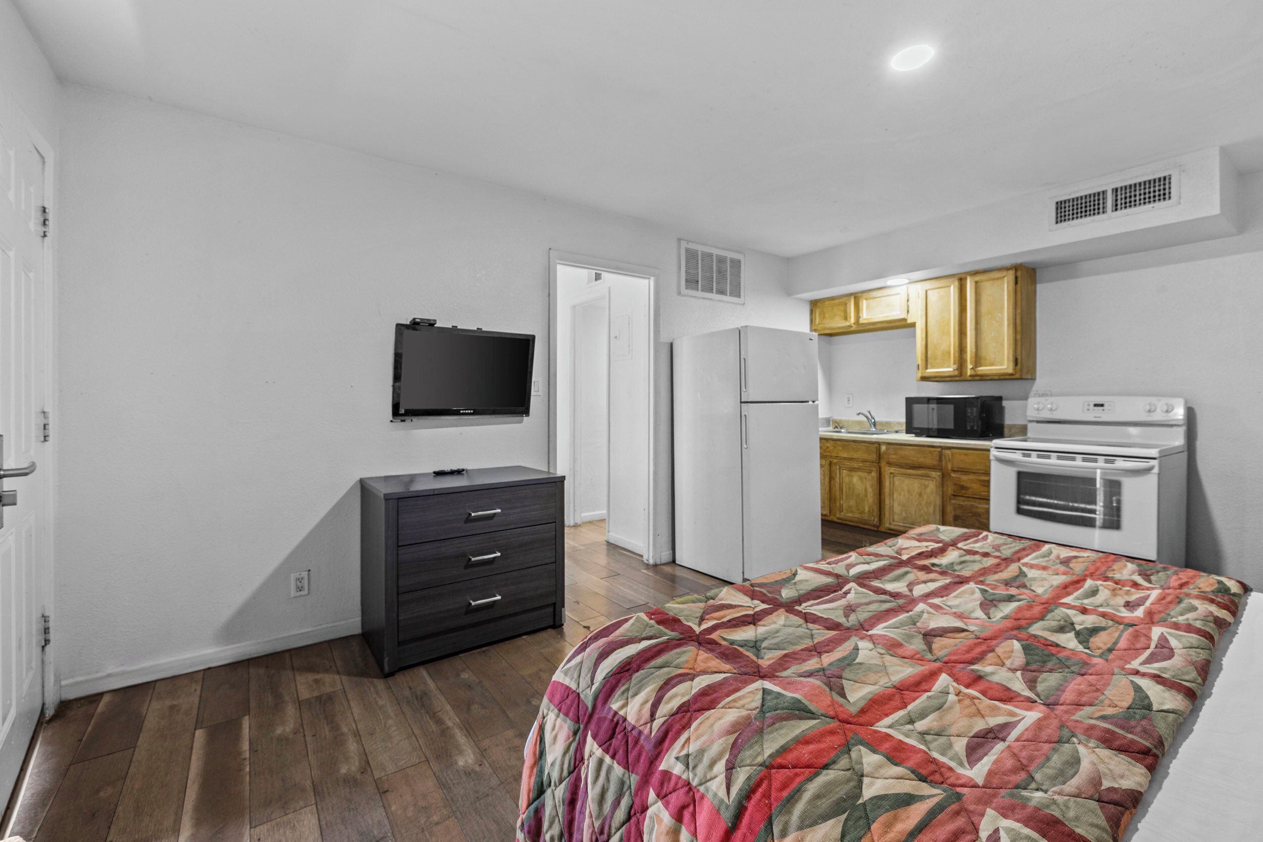 A bedroom with a bed, a dresser, a television, and a kitchenette with a microwave and stove.