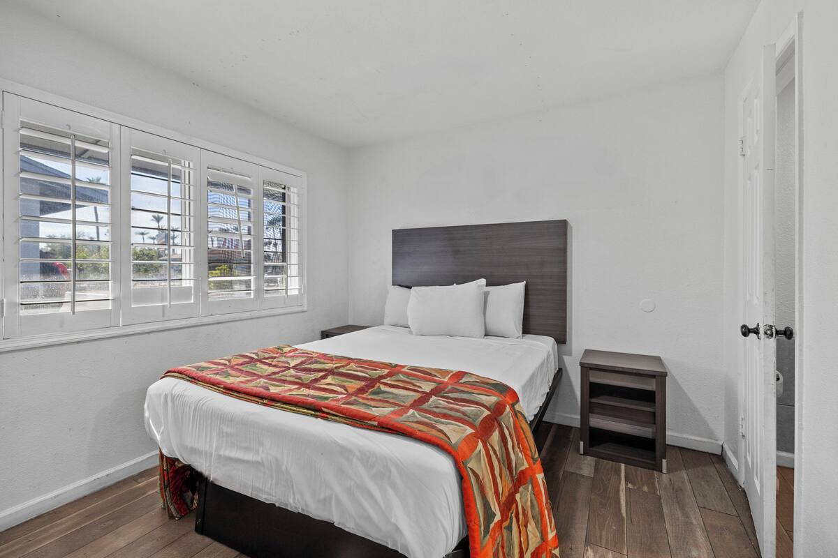 A bedroom with a bed, a nightstand, a window with blinds, and a door.