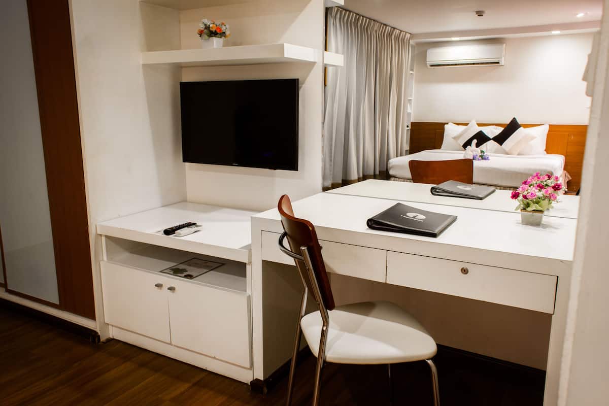 Una camera d'albergo con una televisione a schermo piatto montata a parete, una scrivania con un portatile, una sedia e un letto con biancheria da letto bianca e cuscini decorativi.