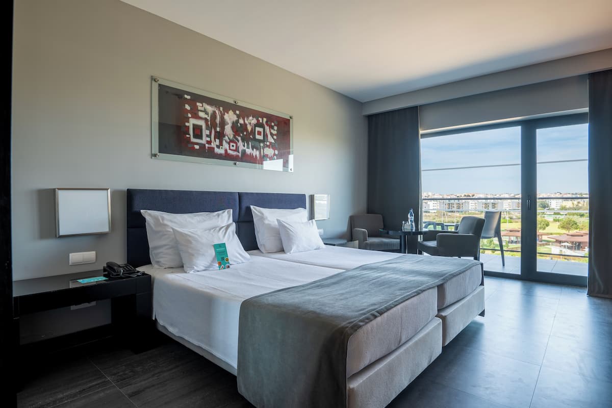 A modern hotel room with a large bed, a desk, and a chair. There is a large window with a view of the city.