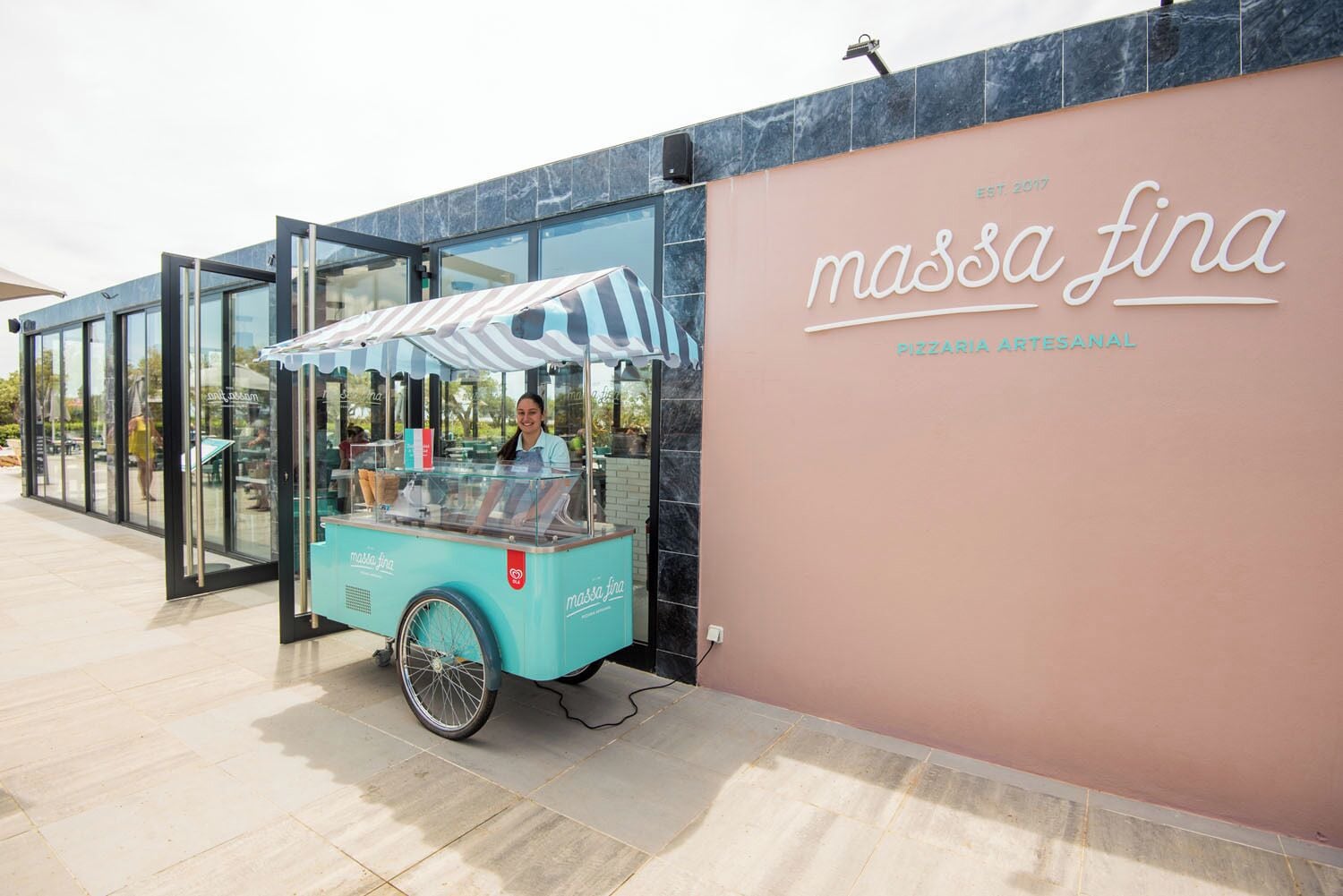 A food cart with a striped canopy labeled "massa fina" in front of a building with a pink exterior and a glass door.