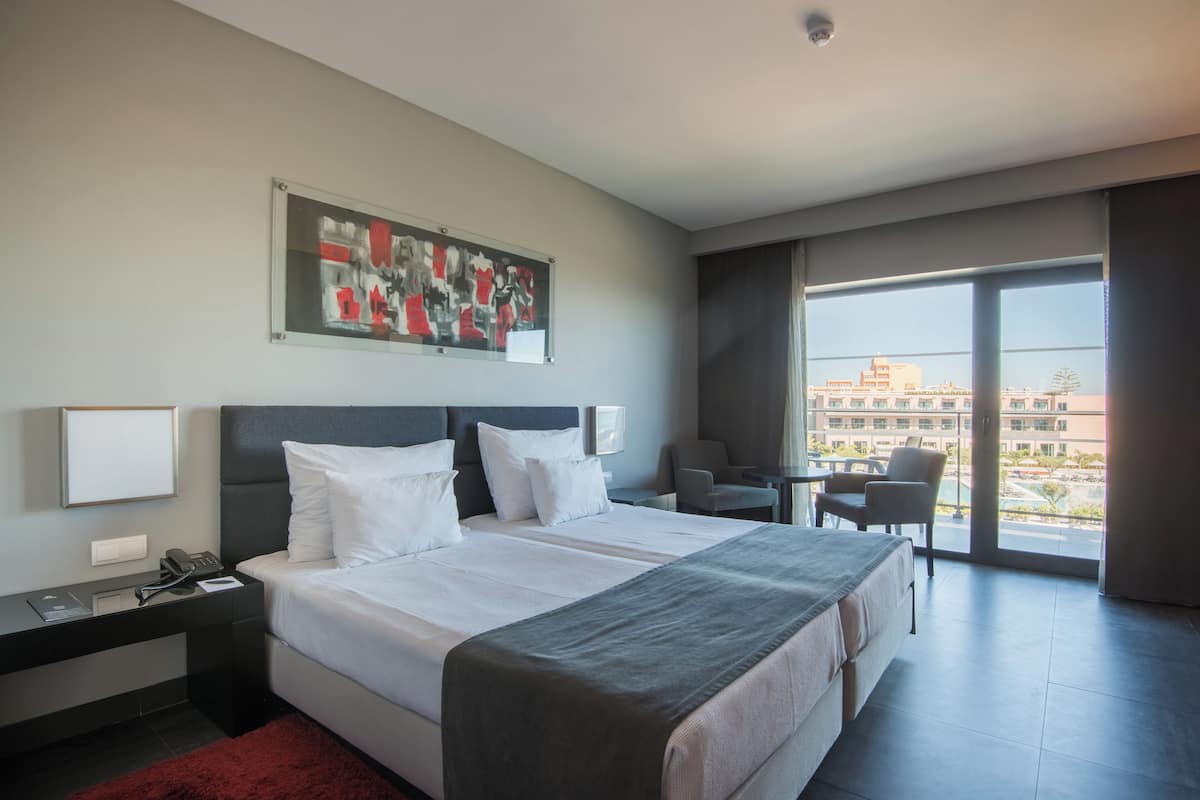 A modern hotel room with a large bed, a desk with a phone, a chair, and a view of the city through large windows.