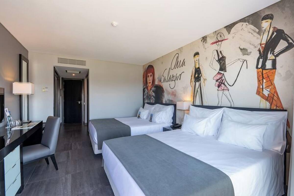 A hotel room with two beds, a desk, a chair, and a mural on the wall.