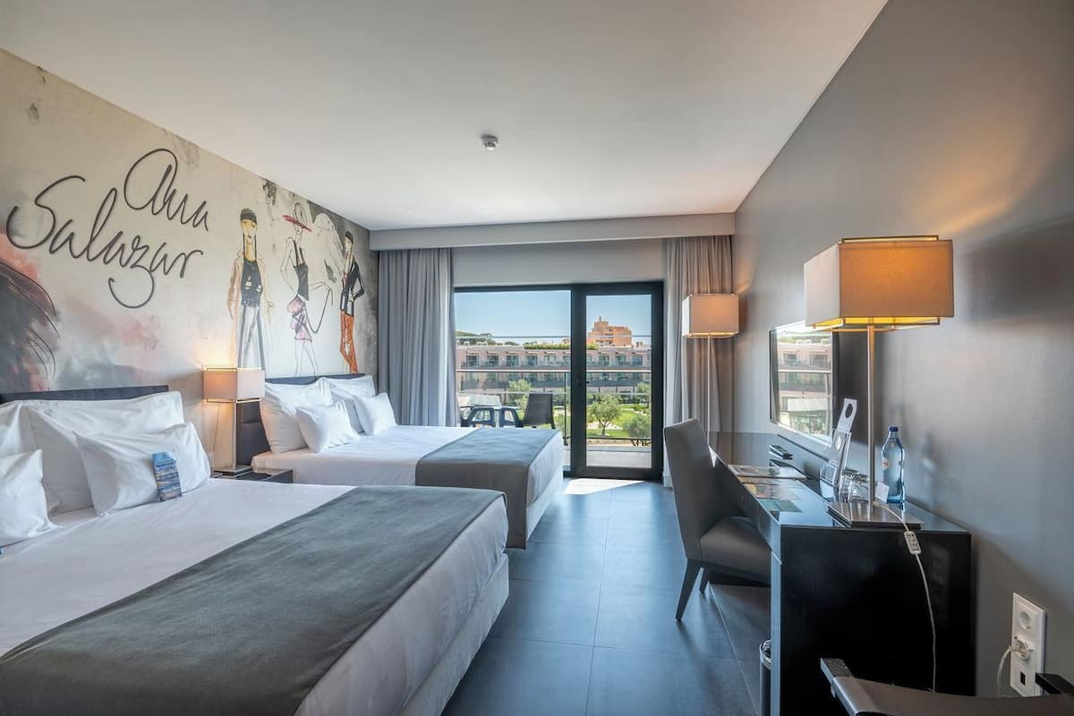 A hotel room with two beds, a desk with a chair, a laptop, a water bottle, and a view of buildings through the window.