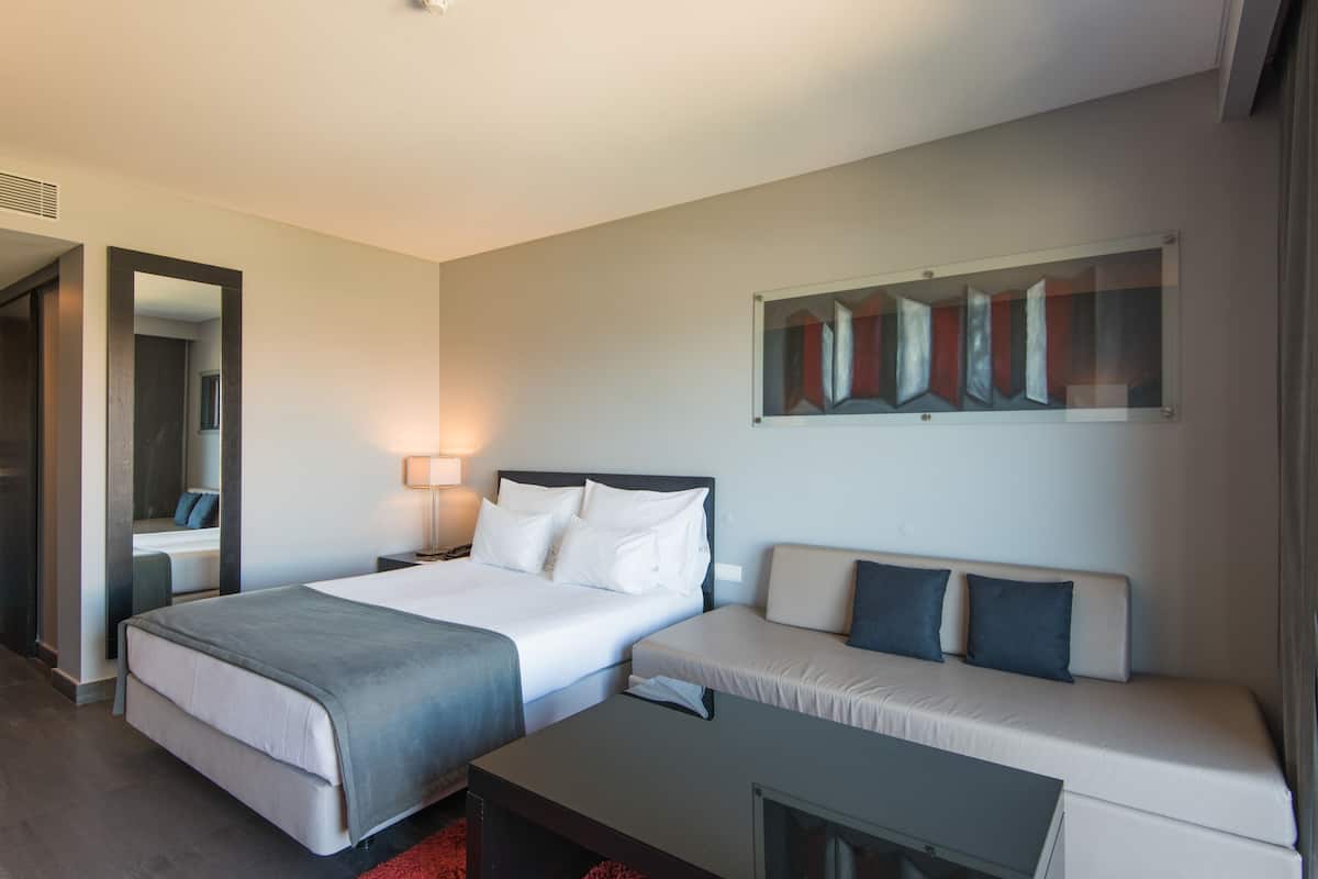 A hotel room with a bed, a sofa, a desk, and a painting on the wall.