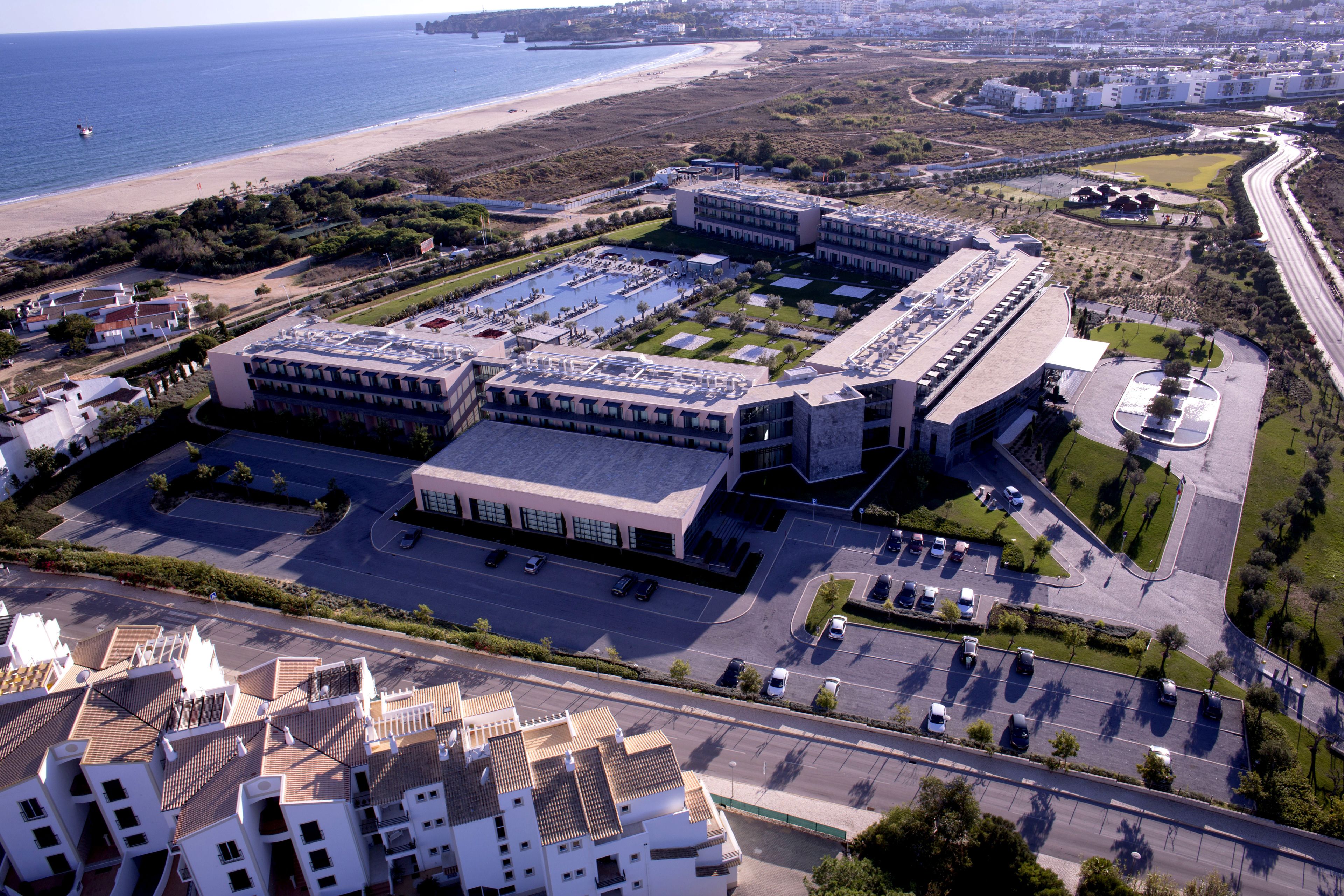 Vista aérea de un complejo moderno de edificios con varios estacionamientos y una playa al fondo.