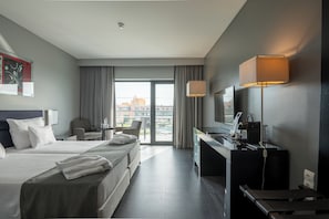 A hotel room with two beds, a desk, a chair, a TV, and a view of the cityscape.
