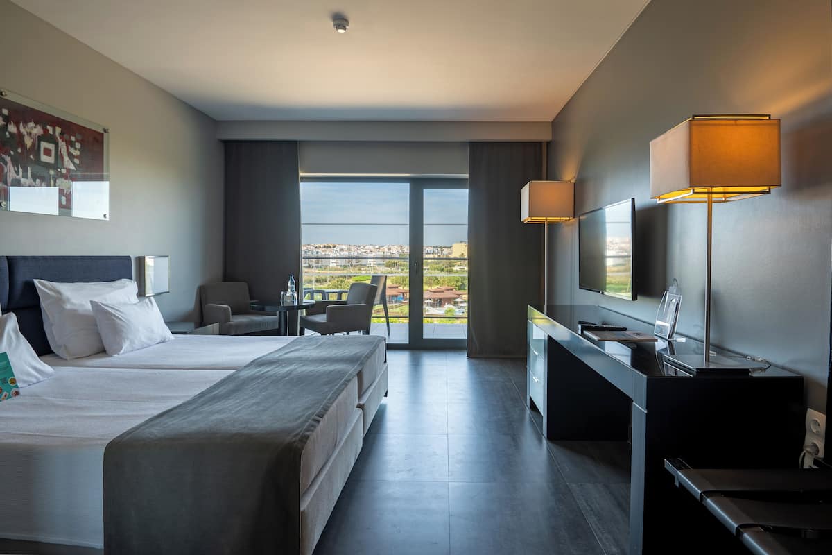 A hotel room with two beds, a desk, a TV, and a large window offering a city view.