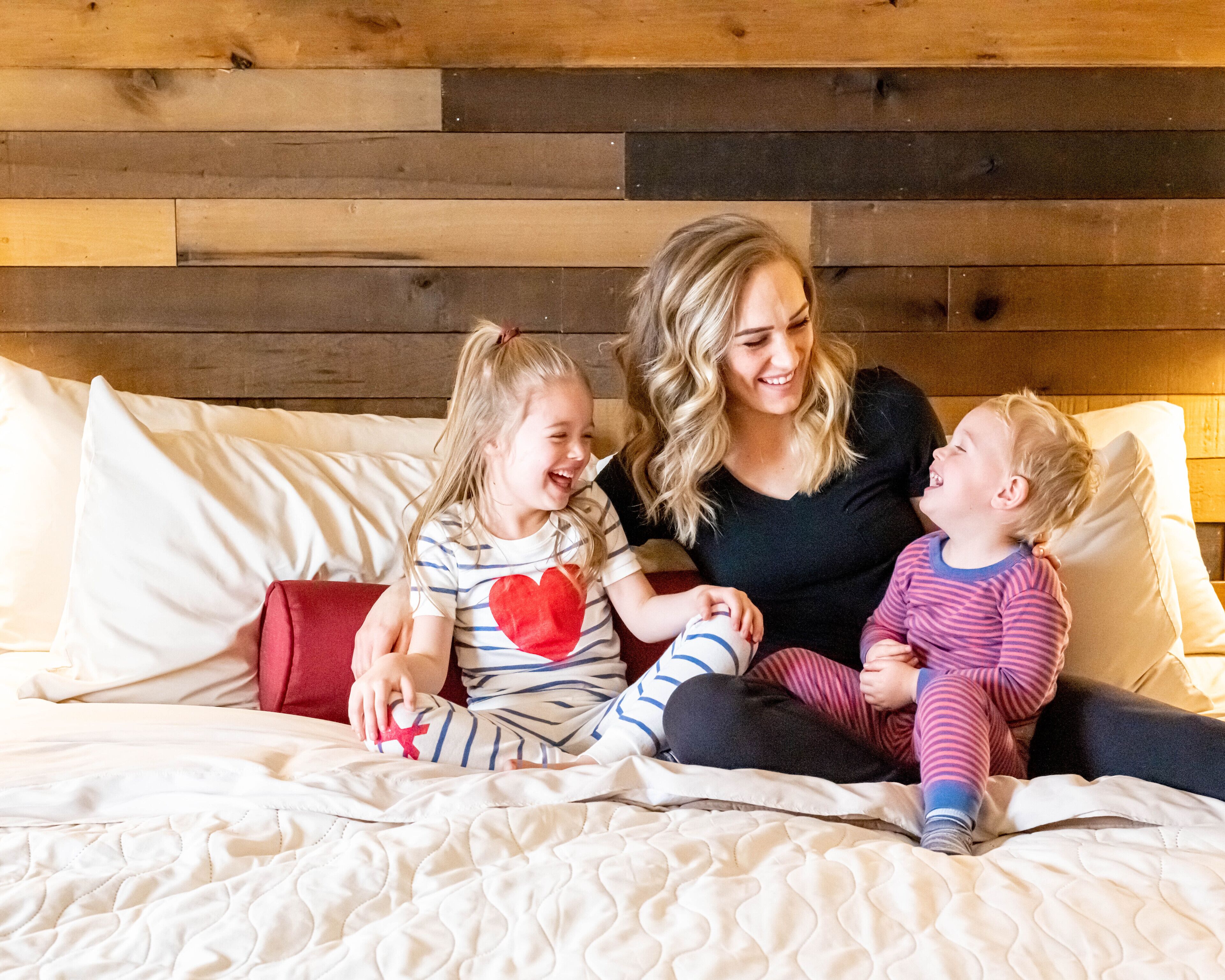 A family of three on a bed with a quilted white cover, wooden headboard, and white pillows.