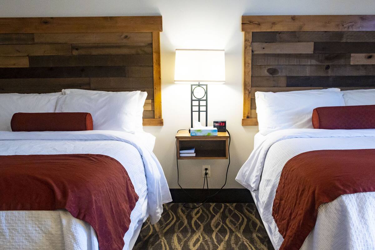 A hotel room with two beds, each with white linens and brown headboards. There is a nightstand with a clock and a lamp between the beds. The room has a wooden headboard design and a patterned carpet.