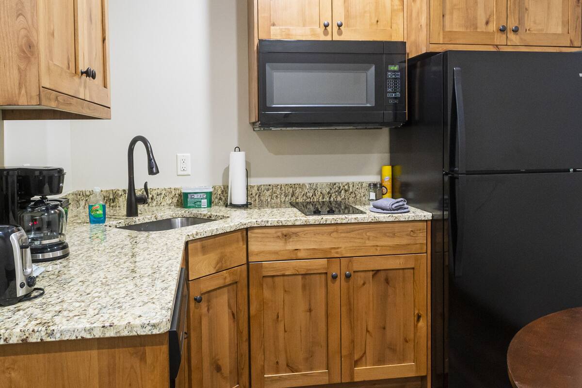 A kitchen with wooden cabinets, a black microwave, a black refrigerator, a granite countertop, a stainless steel sink, and a coffee maker.