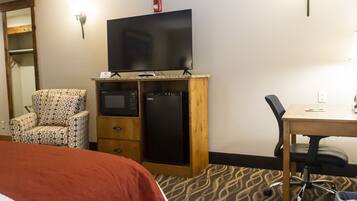 Habitación de hotel con cama, escritorio de madera, silla, microondas, refrigerador y televisor sobre un mueble de madera.