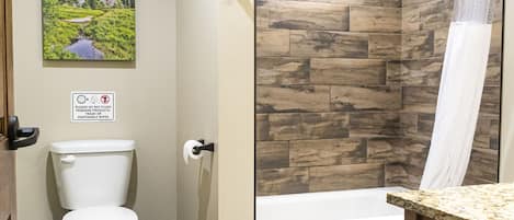 A bathroom with a toilet, a shower with a wooden wall, a sink with a marble countertop, and a sign prohibiting flushing feminine products.