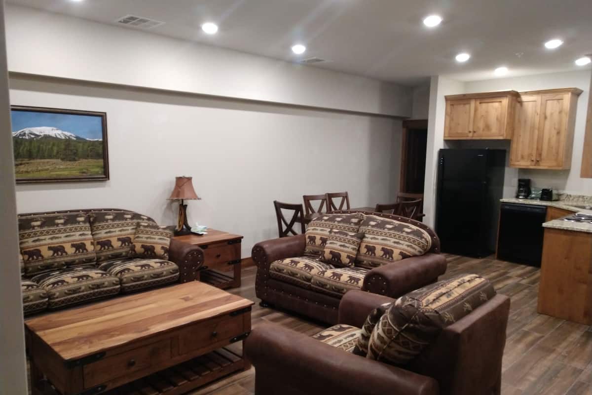 A living room with a sofa set, a wooden coffee table, and a lamp. There is a kitchen area with wooden cabinets and a refrigerator.