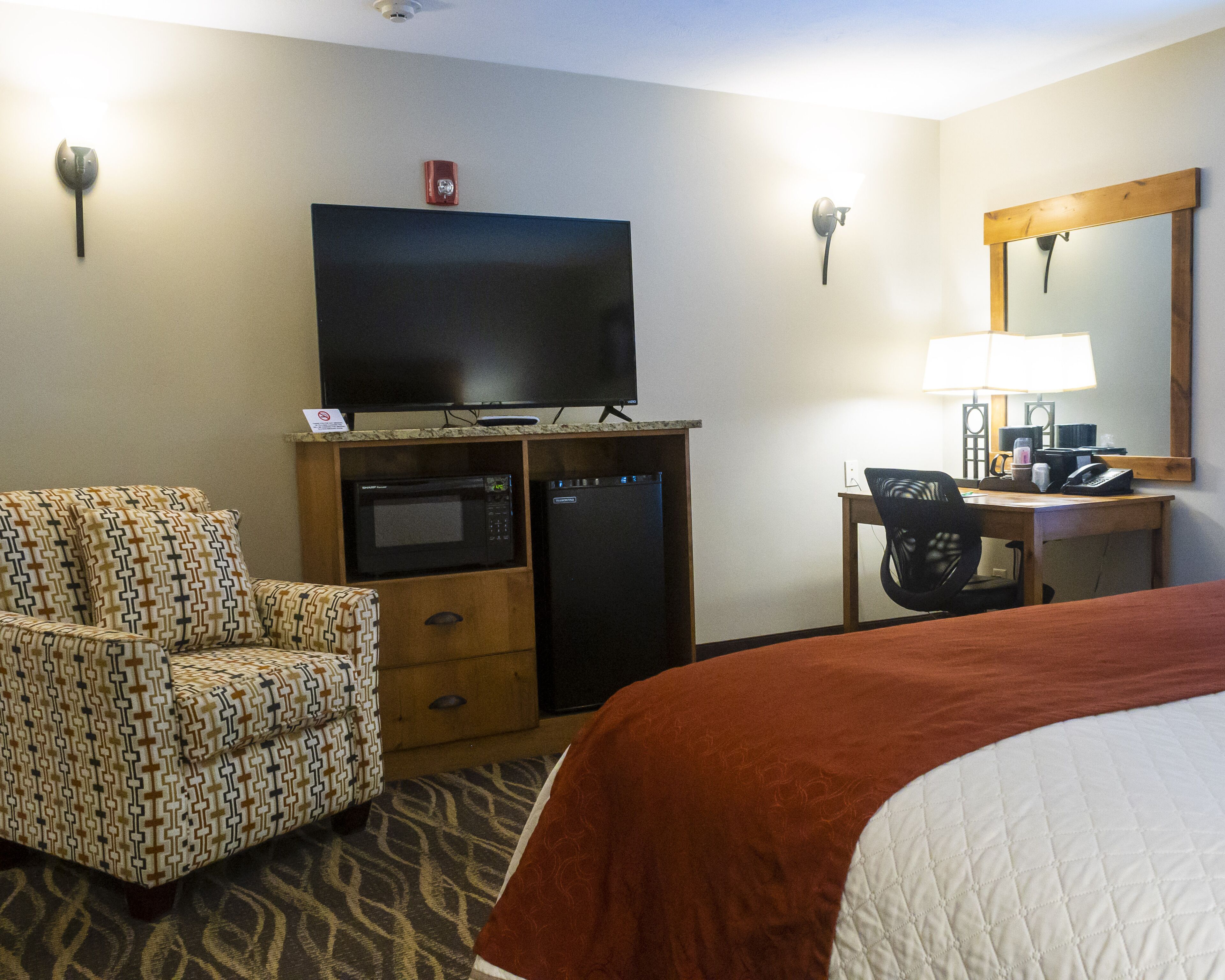 A hotel room with a bed, a television, a microwave, a refrigerator, a desk with a chair, and a mirror.