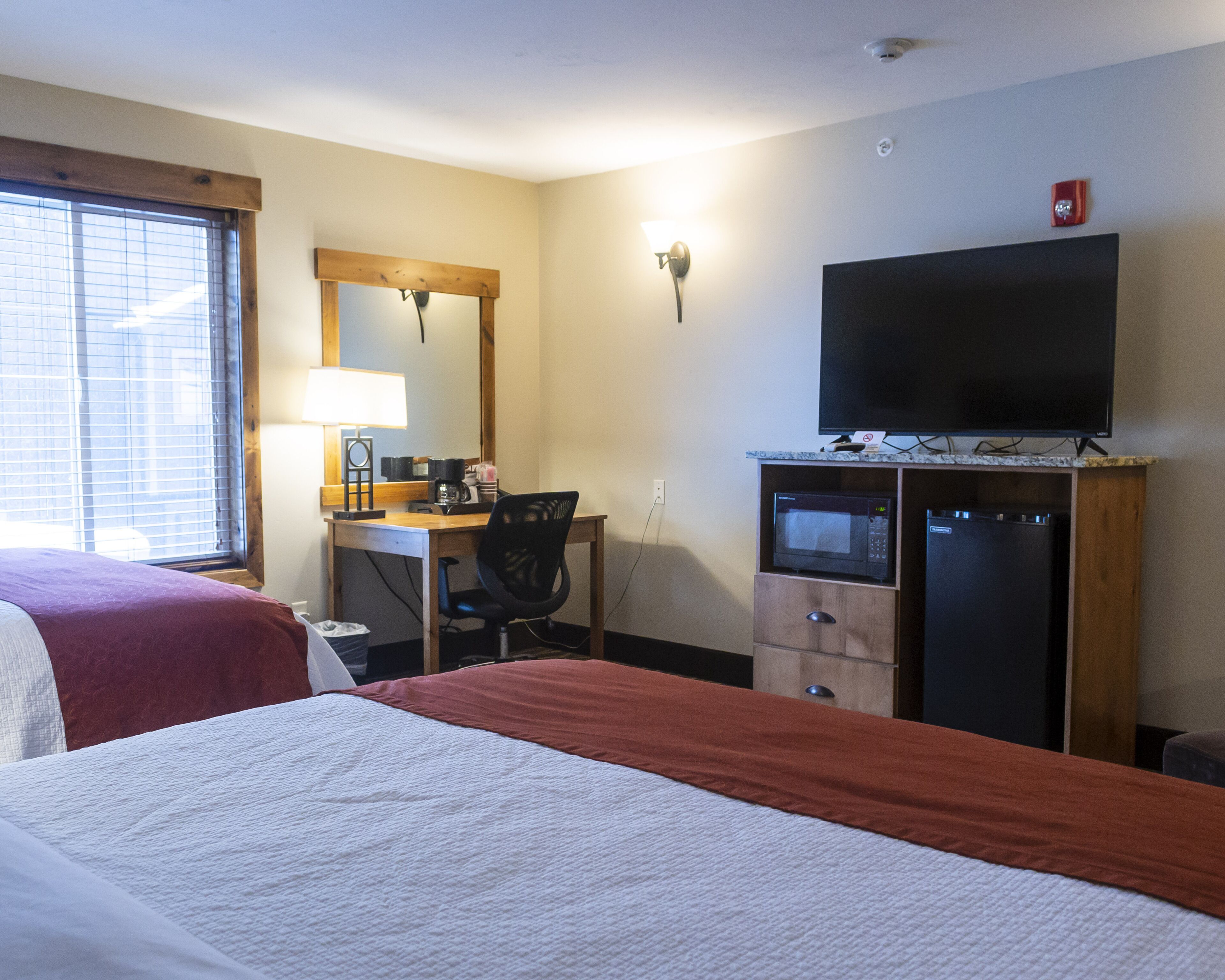 A hotel room with two beds, a desk with a chair, a microwave, a refrigerator, a television, and a window with blinds.