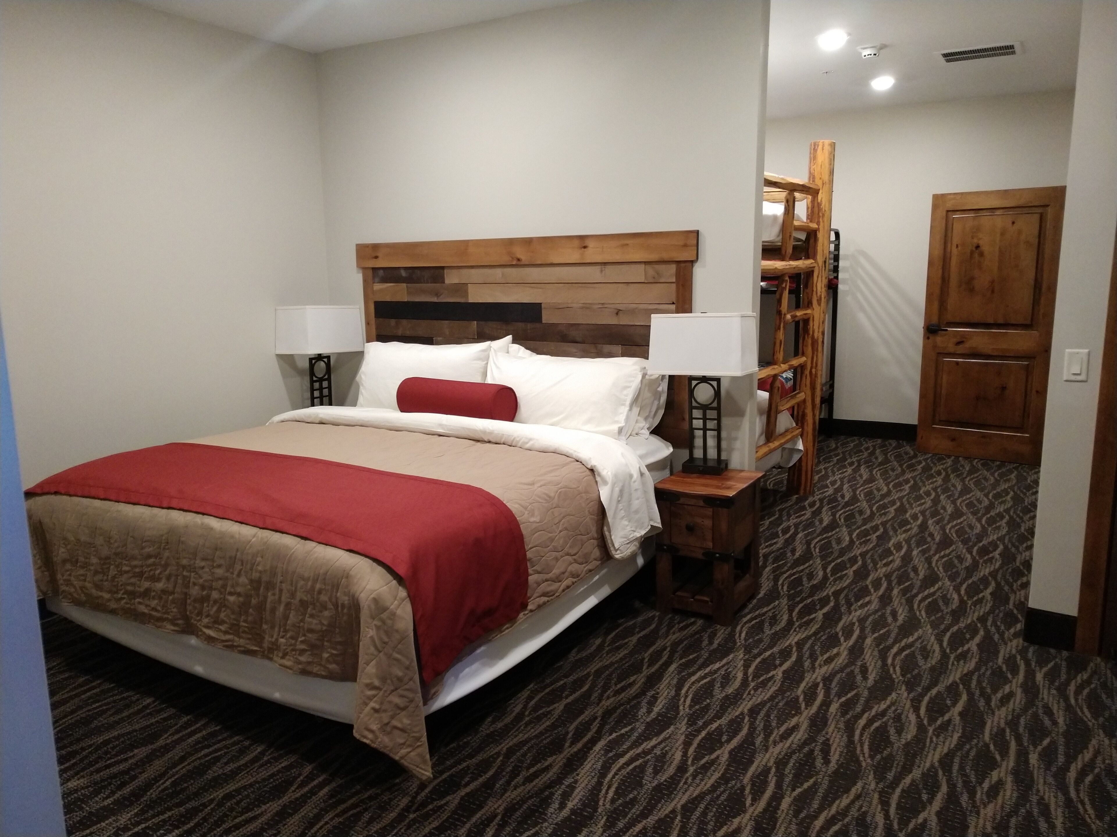 A hotel room with a wooden headboard, a bed with a red and beige bedspread, two bedside lamps, a wooden nightstand, and a patterned carpet.