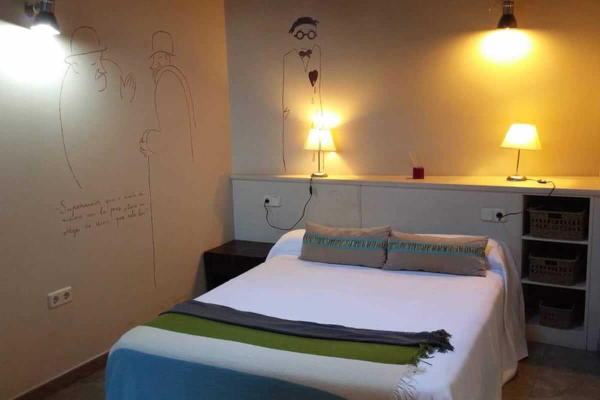 A bedroom with a bed, bedside lamps, a wall with sketches, and a shelf with baskets.
