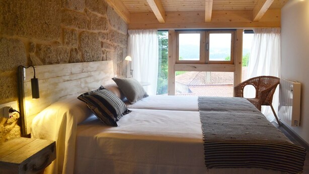 Een slaapkamer met een stenen muur, houten balken in het plafond, een groot bed met kussens, een nachtkastje, een stoel en uitzicht op het landschap.