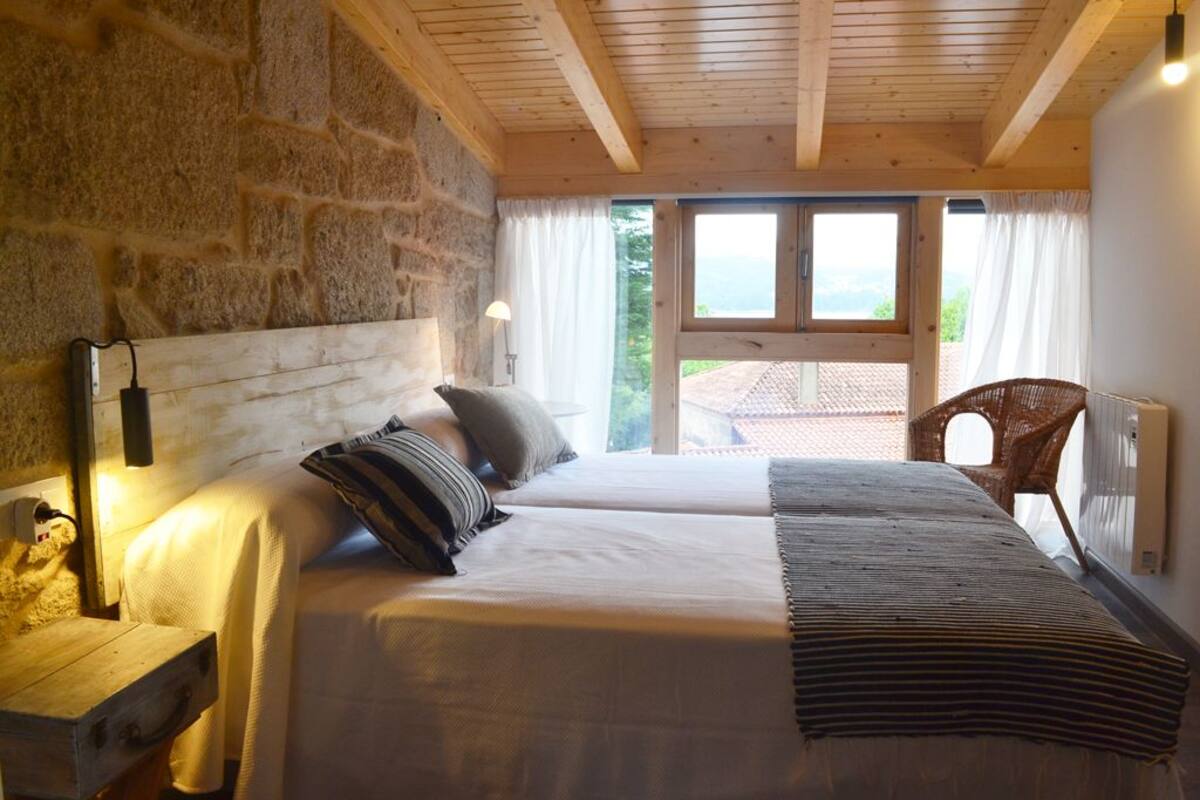 A bedroom with a stone wall, wooden ceiling beams, a large bed with pillows, a nightstand, a chair, and a view of the outdoors.