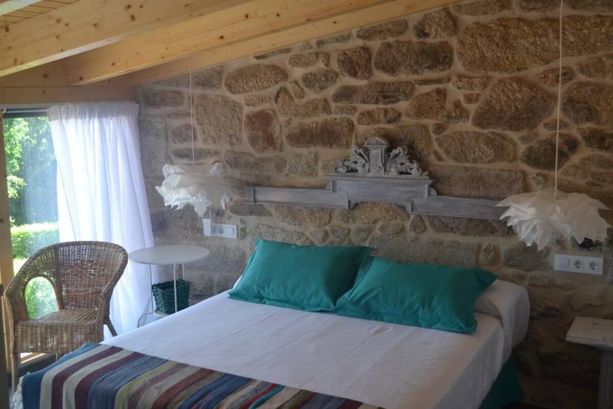 A bedroom with a stone wall, a bed with teal pillows, a wicker chair, and a small table.
