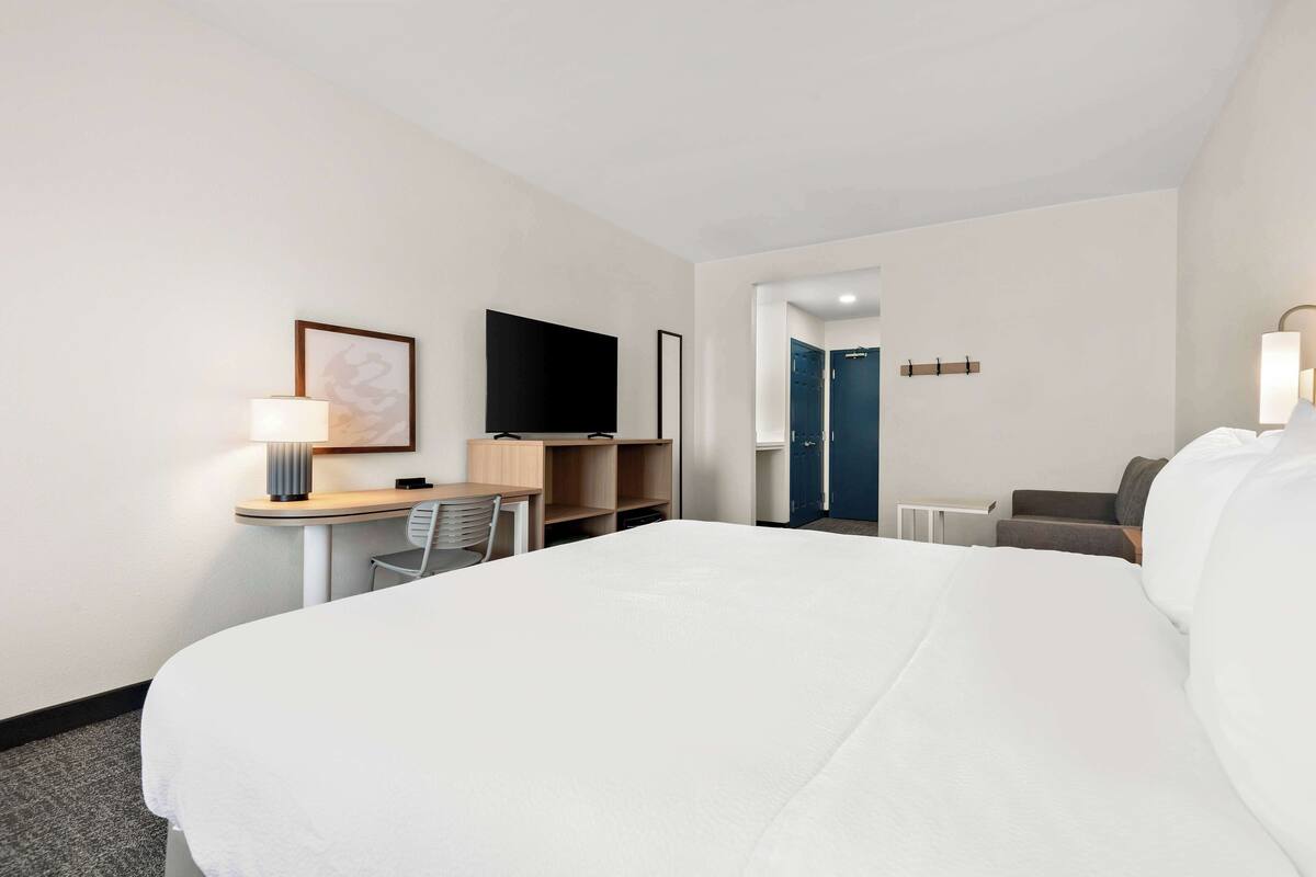 A hotel room with a large bed, a desk with a chair, a flat-screen TV, and a door leading to another room.