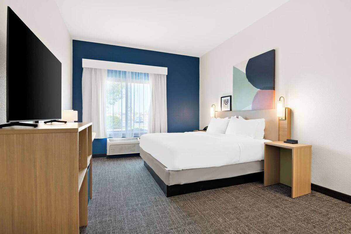 A hotel room with a bed, a desk, a television, a window with curtains, and a wall-mounted light fixture.