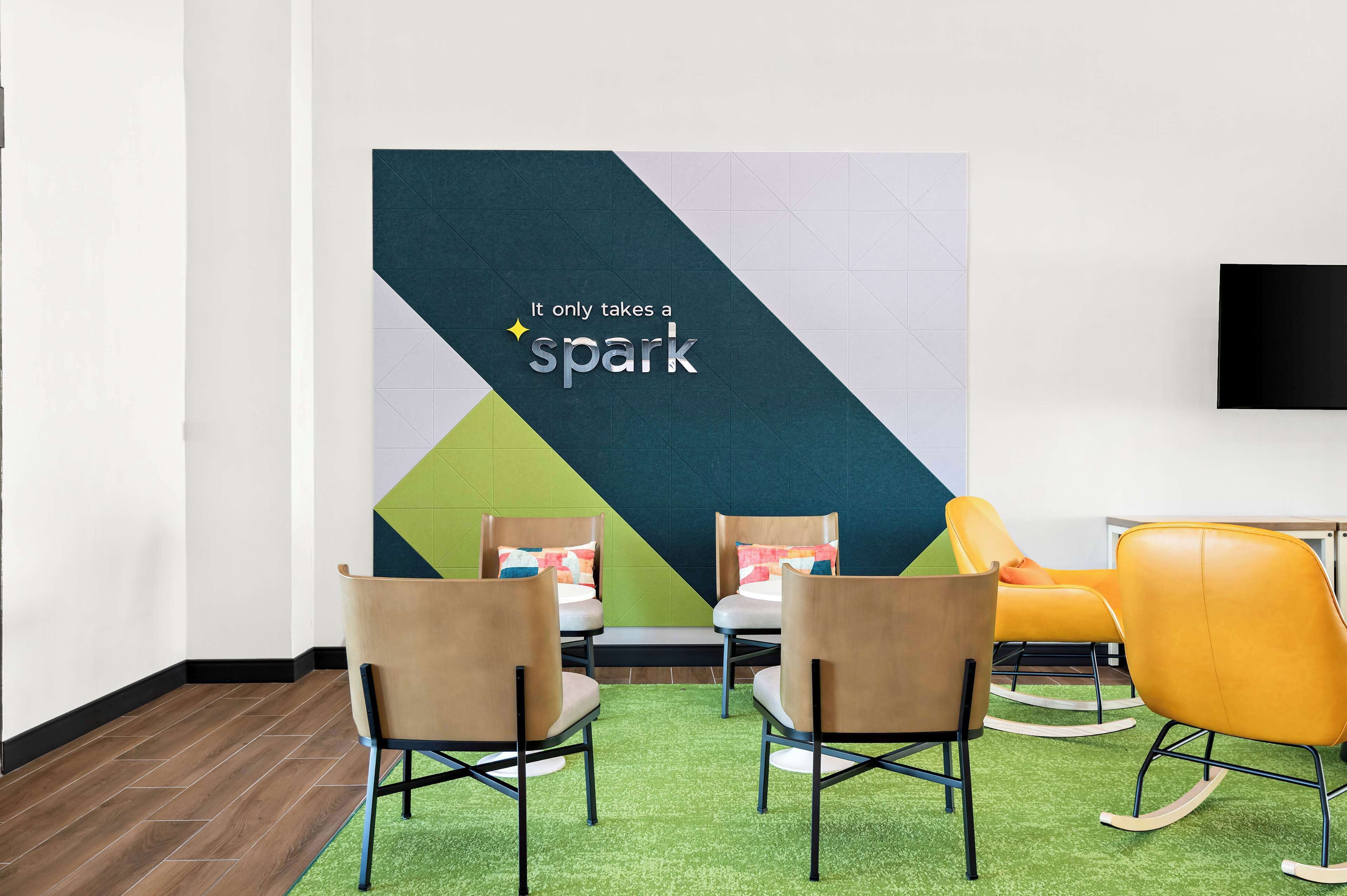 Una habitación con dos sillas marrones, una mecedora amarilla y una alfombra verde. Se ve una pared grande con el texto 'It only takes a spark'.