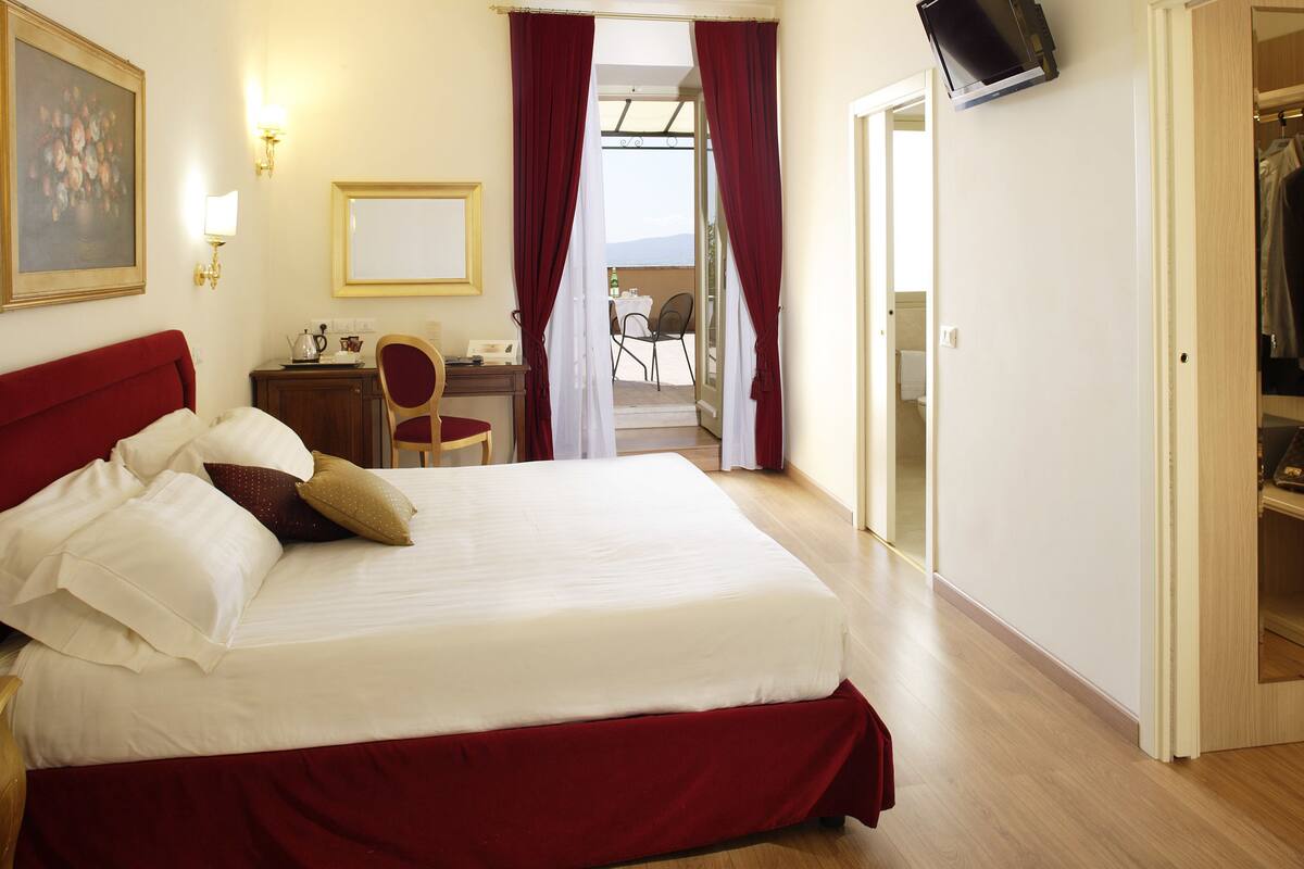 A hotel room with a large bed, a desk with a chair, a TV mounted on the wall, and a view of the outdoors through a door.