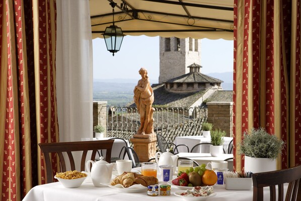 A breakfast table with croissants, fruit, and beverages, overlooking a scenic view with a statue and castle. - Giotto Hotel & Spa (Assisi)