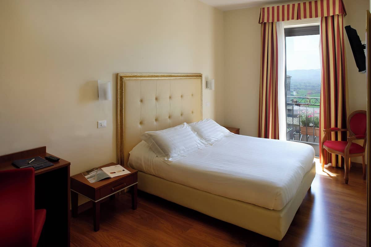 A hotel room with a large bed, a desk with a chair, a small table, and a window with curtains.