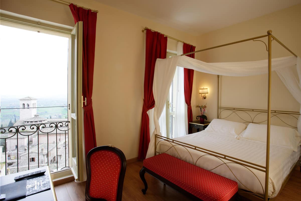 A four-poster bed with a canopy, a red upholstered bench, a chair, a bedside table with a lamp, and a view of buildings through a window with red curtains.