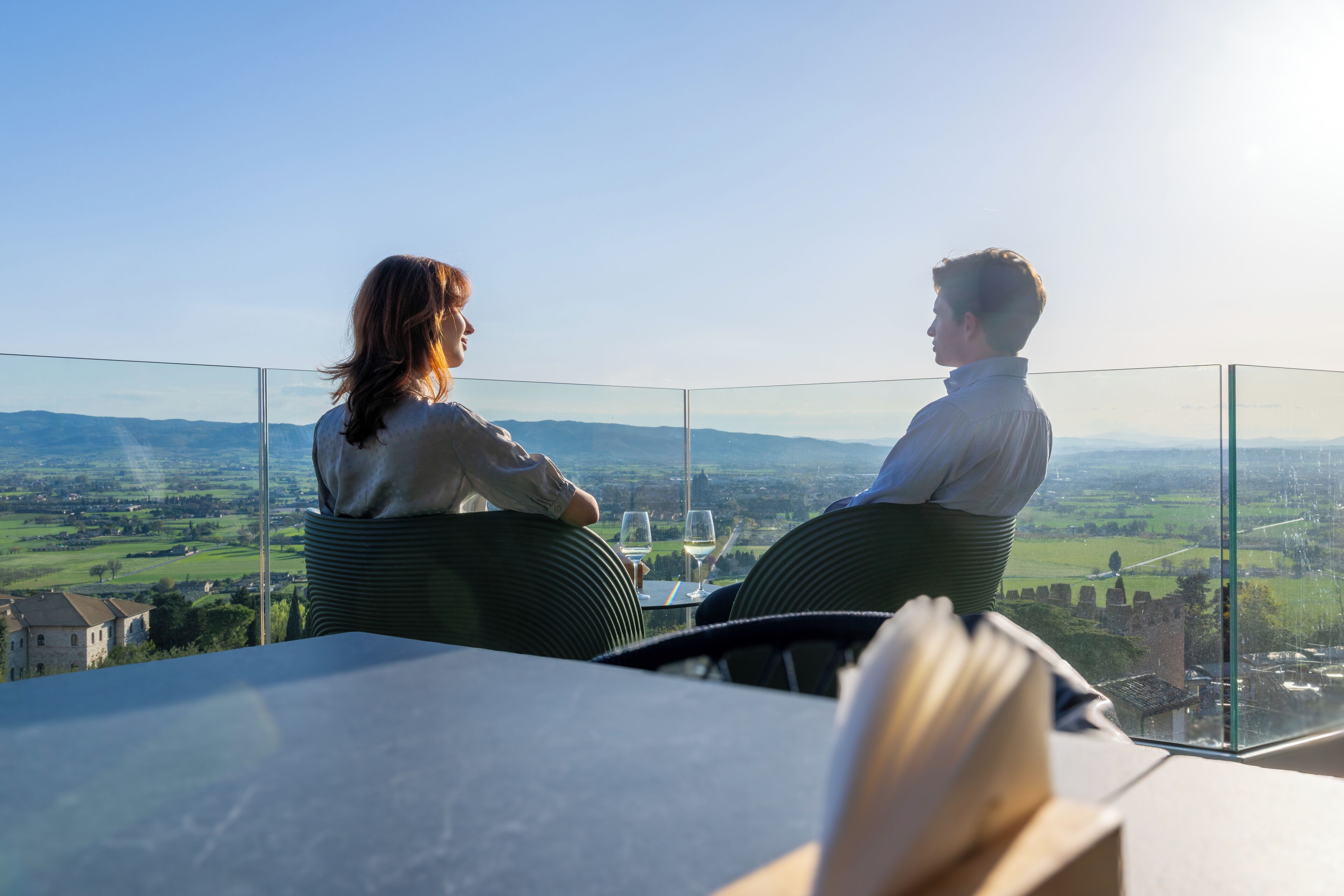 Two people seated at a table with a glass partition, overlooking a scenic landscape with fields and distant buildings.
