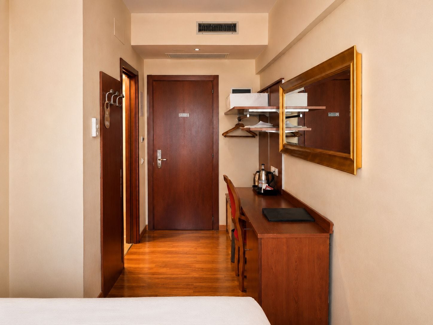 A hotel room with a wooden floor, a bed, a desk with a mirror, and a wardrobe.
