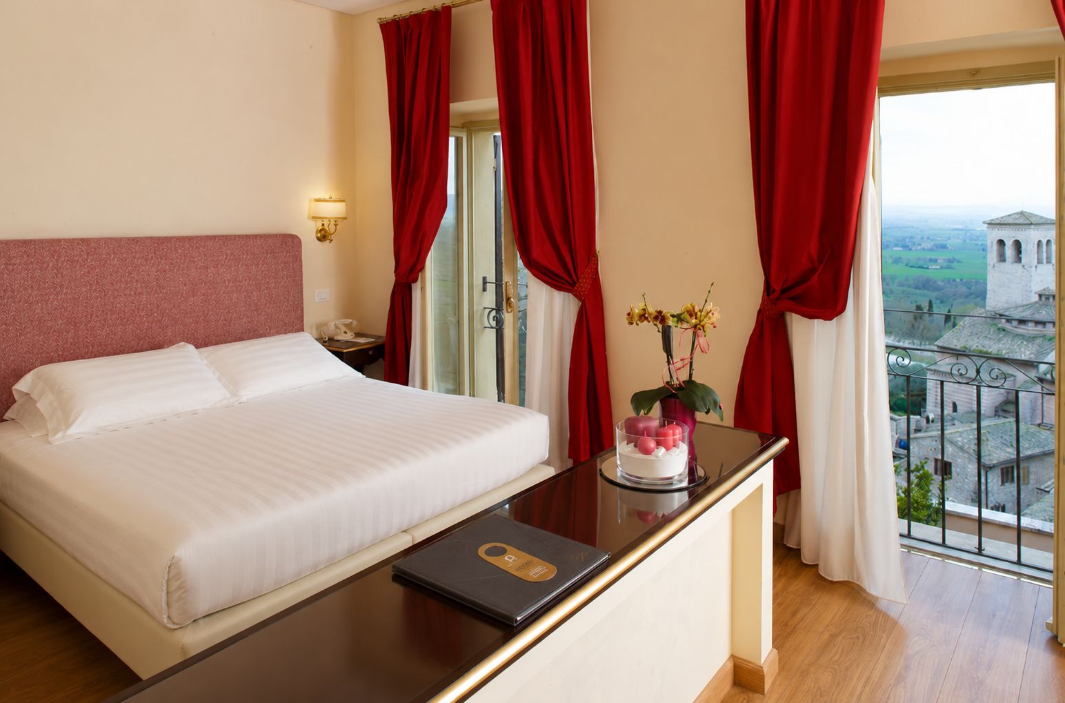 A hotel room with a bed, a bedside table with a laptop, a vase with flowers, and a view of a building through the window.