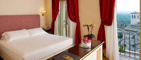 A hotel room with a bed, a bedside table with a laptop, a vase with flowers, and a view of a building through the window.