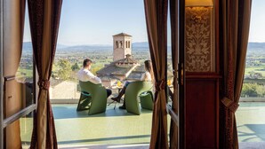 A balcony with two green chairs and a table, overlooking a landscape with a tower and distant buildings. - Giotto Hotel & Spa (Assisi)