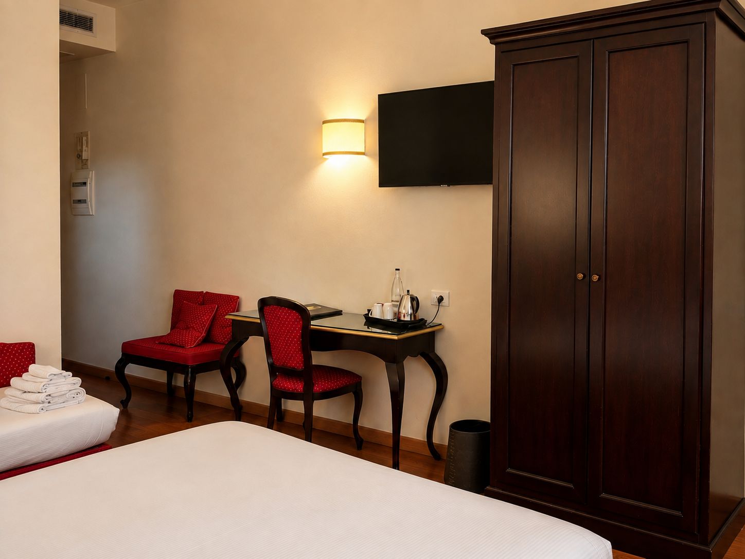 Habitación de hotel con cama, dos sillones tapizados en rojo, una mesita con tetera, televisión montada en la pared y un armario de madera oscura.