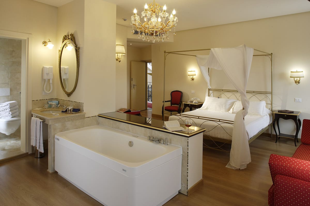 A hotel room with a large bathtub, a four-poster bed, a sitting area with a red chair, and a vanity with a mirror and sink.