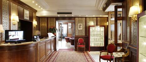A hotel lobby with a reception desk, a red chair, a bookshelf, and a patterned carpet.