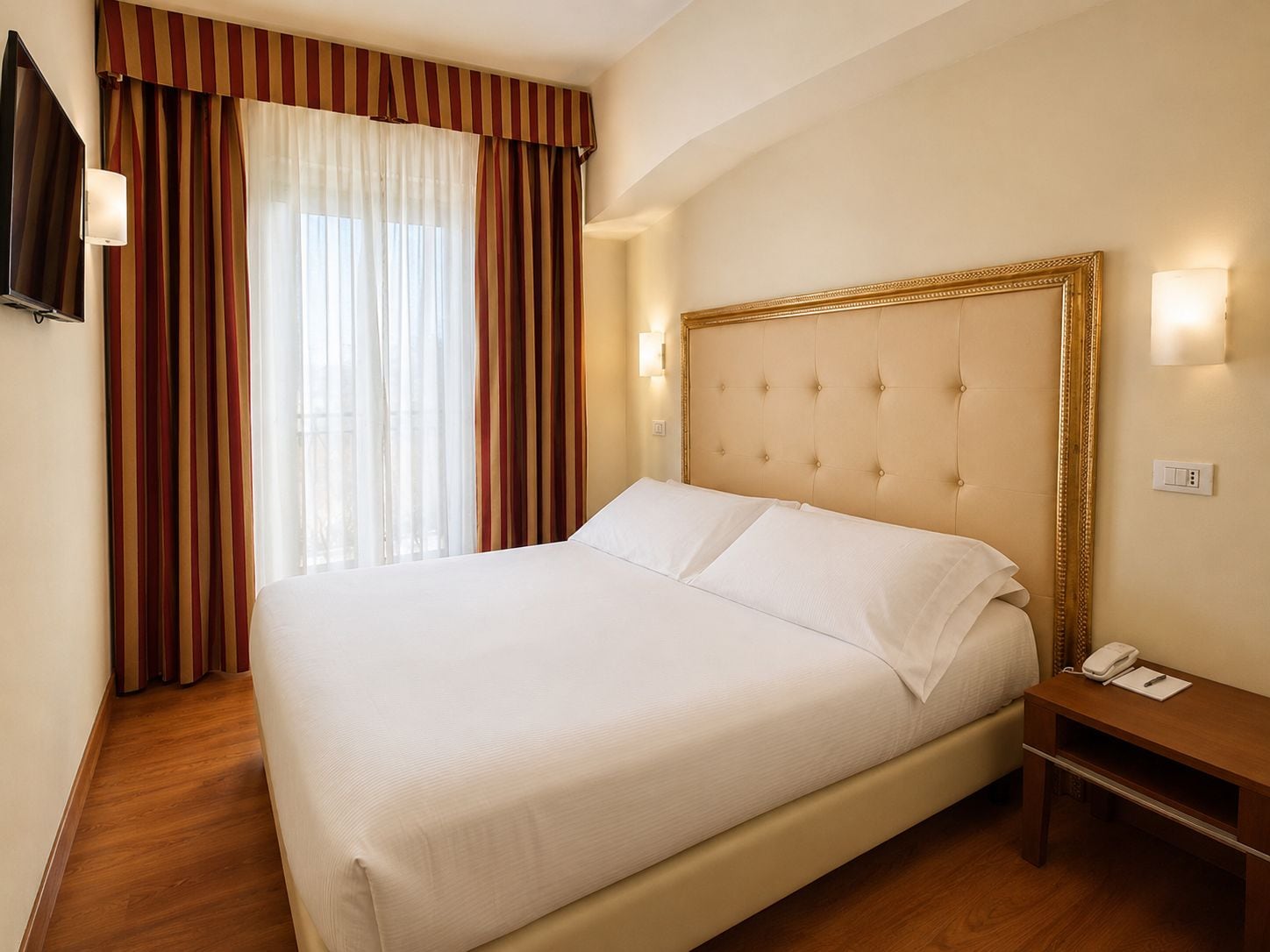 Ein Hotelzimmer mit einem großen Bett, einem Nachttisch mit Lampe und einem an der Wand befestigten Fernseher.